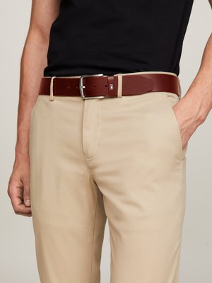 Cinturón de cuero marrón con una hebilla de metal plateado, llevado sobre pantalones beiges. El cinturón tiene una forma rectangular y un diseño minimalista.