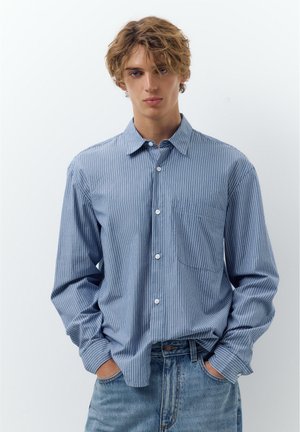 Young man with curly blond hair wearing a blue and white striped button-down shirt and light blue jeans with hands in pockets.