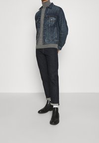 Mörk denimjacka över en grå polotröja, parat med mörka denimjeans och svarta ankelskor. Har klassiska fickor och uppvikta ärmar.