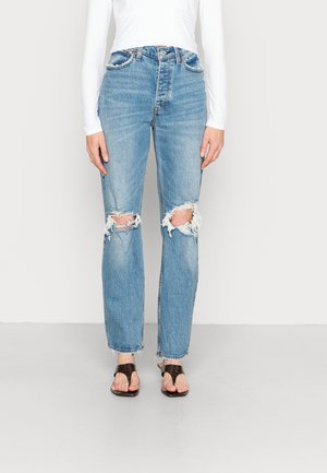 Jeans de ganga de tom azul claro com joelhos rasgados, design de perna reta, bainhas desfiadas, textura desgastada, emparelhados com sandálias planas castanhas.