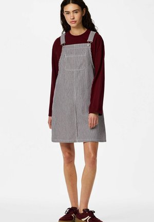 Kobieta ubrana w bordową bluzkę z długim rękawem pod białą i czarną pasiastą sukienkę z kieszenią z przodu oraz bordowe trampki, stoi nieruchomo.