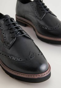 Čierne kožené brogues s perforovanými detailmi, hrubými hnedými okrajmi na podrážkach a čiernymi tkaničkami na svetlom povrchu.
