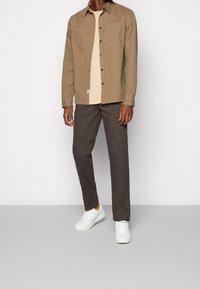 Beżowa koszulka z dwoma przednimi kieszeniami, kremowa koszulka pod spodem, ciemnobrązowe spodnie i białe trampki, tworzące neutralną, warstwową stylizację.