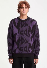 Lilla og svart mønstret genser med rund hals, lange ermer og ribbestrikkede mansjetter, laget av mykt strikket stoff.