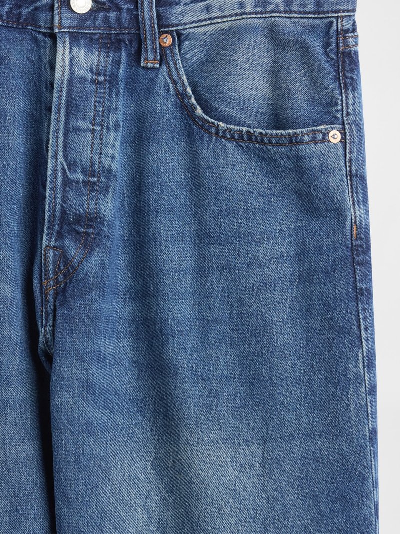 Nahaufnahme von blauen Jeans, die die Vordertasche, den Gürtelschlaufe, den Metallknopf und die orangefarbenen Nähte zeigen.
