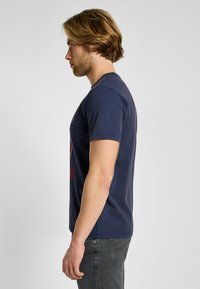 Camiseta de algodón azul marino con cuello redondo y mangas cortas, con letras rojas en el frente. Combinada con pantalones grises.