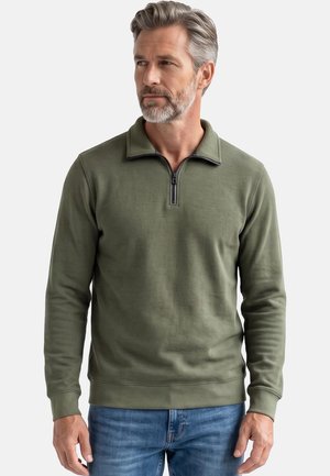 Uomo con capelli grigi e barba indossa un maglione verde oliva con cerniera e jeans blu, guardando di lato contro uno sfondo semplice.