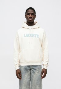 Camisola com capuz creme com um logótipo "LACOSTE" macio e felpudo em cores pastel, um bolso canguru e cordões, combinado com jeans de lavagem clara.