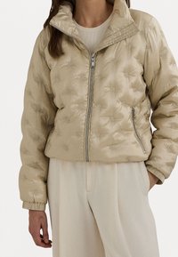 Beżowa kurtka puchowa z zamkiem z przodu, wysokim kołnierzem i misternym, tłoczonym wzorem logo. Posiada ściągacze na mankietach i dole, zapewniające dopasowanie.