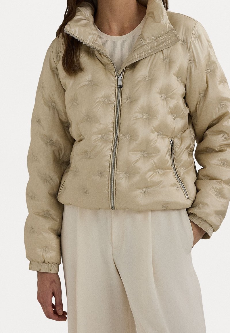 Beżowa kurtka puchowa z zamkiem z przodu, wysokim kołnierzem i misternym, tłoczonym wzorem logo. Posiada ściągacze na mankietach i dole, zapewniające dopasowanie.