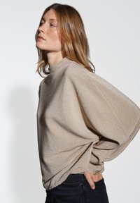 Donna con capelli lunghi fino alle spalle, che indossa un maglione beige a coste con maniche voluminose e pantaloni scuri, in piedi di lato su uno sfondo neutro.