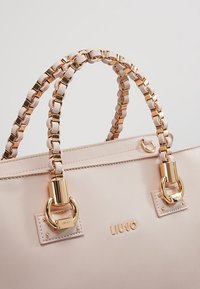 Sac à main beige avec des poignées en chaîne dorée et des ferrures, avec le nom de la marque "Liu Jo" en lettres dorées sur le devant.