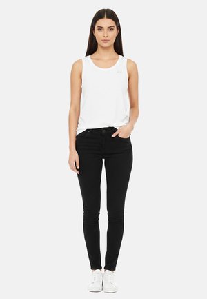 Donna con capelli lunghi e scuri che indossa una canotta bianca senza maniche, jeans skinny neri e scarpe da ginnastica bianche, in piedi con una mano in tasca.