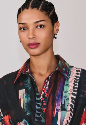 Naine, kellel on punutud juuksed ja hõbedased rõngaskõrvarõngad, kannab mitmevärvilist abstraktse mustriga kraega särki, taust on ühtlane.