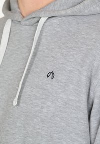 Harmaa huppari pehmeästä kankaasta, jossa on etutasku, naru hupussa ja pieni musta brodeerattu logo rinnassa.