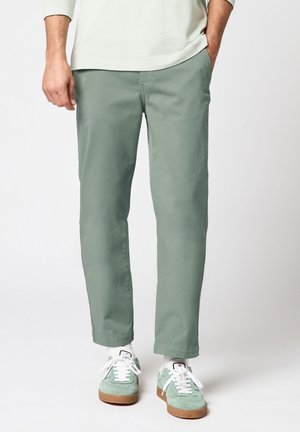 Homme portant un pantalon chino vert sauge avec des chaussettes blanches et des baskets en daim vert clair, debout avec une main dans la poche, sur fond blanc.