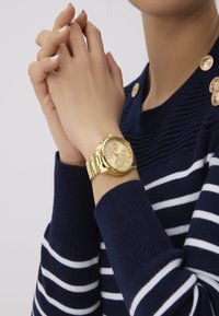 Gul armbåndsur med en tekstureret urskive, store timemarkører og et sammenkoblet metalarmbånd, båret på en hånd mod en marineblå stribet sweater.