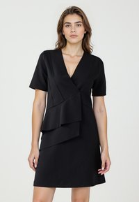 Robe noire à manches courtes, décolleté en V profond et détail de volant asymétrique à l'avant. Fabriquée à partir d'un tissu lisse et extensible.