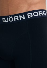 Musta boxershortsi, jossa on laivastonsininen vyötärö, jossa on valkoisella teksti "BJÖRN BORG". Sujuva materiaali ja päältä sopiva muotoilu. Vahvistetut saumat.