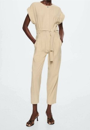 Beżowy kombinezon o dopasowanym kroju, z paskiem w talii, krótkimi rękawami, guziczkami z przodu, kieszeniami oraz czarne sandały na wysokim obcasie z paskami.
