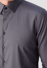 Homme portant une chemise à boutons gris foncé avec le logo "ck" brodé sur le côté gauche de la poitrine.