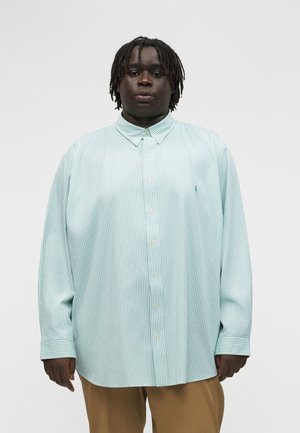 Juodaodis žmogus su trumpais dreideliais, dėvintis šviesiai mėlyną ir baltą dryžuotą per didelį marškinių stilių ir smėlio spalvos kelnes, stovintis ramiai.