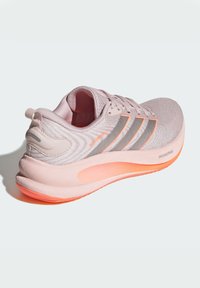 Scarpa atletica rosa chiaro con tomaia in maglia tessuta, tre strisce grigie e suola arancione vivace. Include una linguetta sul retro per indossarla facilmente.