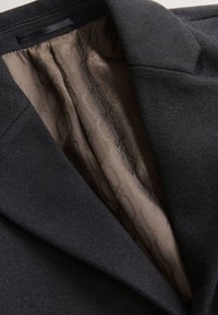 Col de manteau en laine noire avec doublure intérieure en satin beige foncé texturé présentant des motifs géométriques subtils.