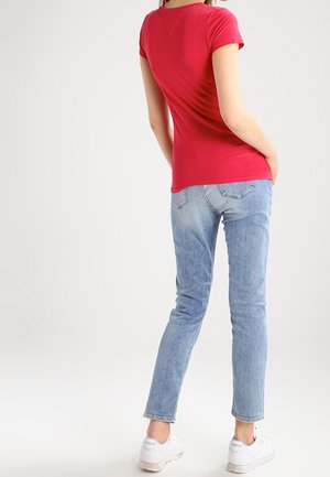 Eine Frau trägt ein rotes Kurzarmshirt, hellblaue Jeans und weiße Sneaker. Sie steht mit den Händen in den Taschen und blickt weg.