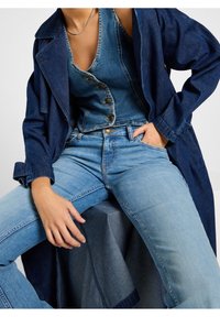 Chaqueta de mezclilla en azul oscuro, superpuesta a un chaleco de mezclilla azul con detalles de botones y jeans de tiro alto en azul claro, mostrando varios tonos de mezclilla.