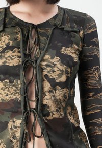 Oberteil mit Camouflage-Muster und V-Ausschnitt, mit Bindebändern vorne. Aus weichem Stoff, hauptsächlich in dunklen Grüntönen und Brauntönen.