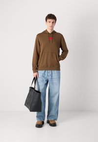 Brun hettegenser med frontlomme, rød logopatch; lyseblå baggy jeans; svart totebag med preget logo; brune sko.