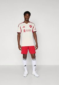 Vitt fotbollströja med röda detaljer, med en logotyp och text. Matchas med röda shorts, vita strumpor med svarta ränder och vita fotbollsskor.