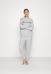 Pilkas sportinis laisvalaikio kostiumas su ilgomis rankovėmis ir kelnėmis, įskaitant žalią "LACOSTE" logotipą krūtinės srityje. Minkšta tekstūra, laisvas siluetas.