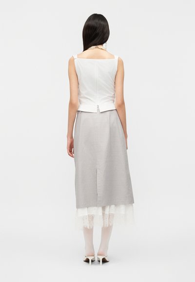 Γυναίκα με μακριά σκουρόχρωμα μαλλιά φοράει λευκό αμάνικο τοπ, γκρι μίντι φούστα με δαντελωτό τελείωμα, διάφανα λευκά καλσόν και λευκά παπούτσια με τακούνι, με την πλάτη στραμμένη.