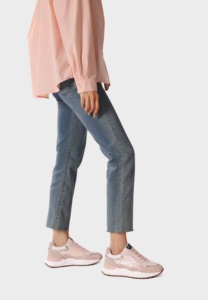 Persona che indossa jeans corti blu, sneaker rosa chiaro e una camicia rosa pallido a maniche lunghe, in piedi con una gamba leggermente piegata.