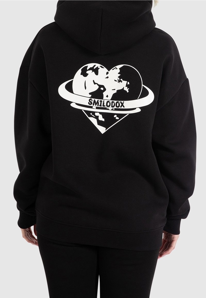 Sweat à capuche noir avec un grand graphisme blanc représentant un cœur entourant un globe, avec le texte "SMILODOX" en évidence. Tissu doux, coupe oversize.