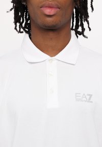 White polo shirt made of cotton, featuring a classic collar, three buttons, and a subtle grey EA7 logo on the left chest.