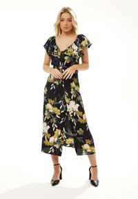 Floral wrap dress in black fabric, featuring yellow and pink blooms, ruffled short sleeves, and a mid-length hem, paired with black heels.