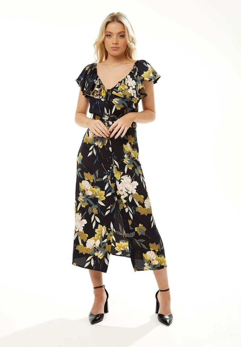 Floral wrap dress in black fabric, featuring yellow and pink blooms, ruffled short sleeves, and a mid-length hem, paired with black heels.