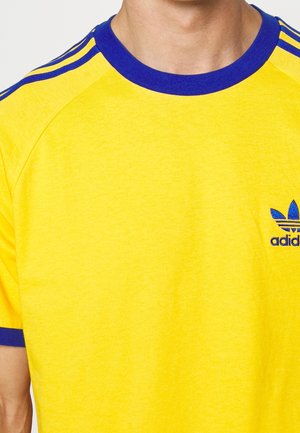 T-shirt jaune Adidas avec col bleu, bordure aux manches et trois bandes sur les épaules, porté par une personne montrée du cou jusqu'au milieu du torse.