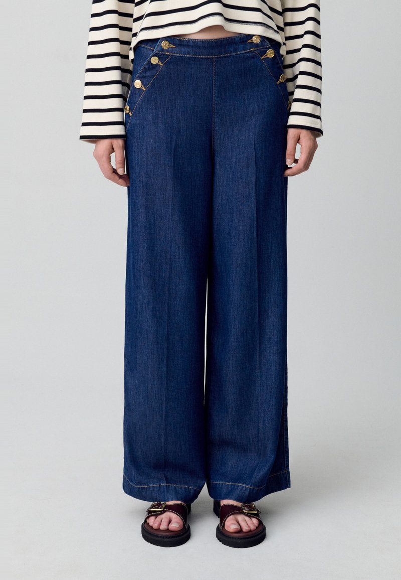 Persona que lleva pantalones de mezclilla oscuros de pierna ancha con botones dorados decorativos, combinados con una camiseta de rayas de manga larga y sandalias oscuras.
