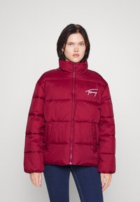 Bordeaux pufferjakke med høy krage, glidelås og Tommy-logo på brystet. Den har to frontlommer og et quiltet design.