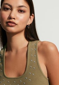 Olivi vihreä tank top, jossa on uritettu pinta, ja hopeanvärisiä koristeita kaula-aukossa ja olkaimissa. Mallilla on korvakorut.