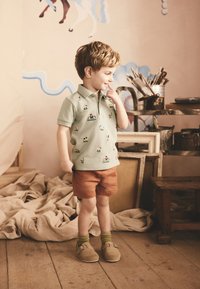 Polo vert pâle avec des motifs brodés colorés, associé à un short rouille. Chaussures marron à boucle et chaussettes vertes. Sol en bois.