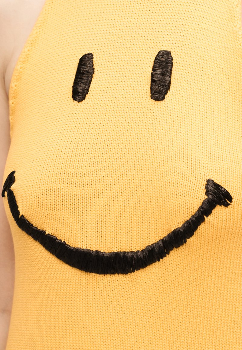 Yellow knit top with a large black embroidered smiley face, featuring oval eyes and a curved smile, worn by a person.