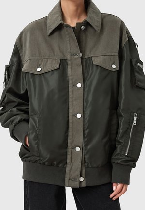 Dark green and olive jacket with snap buttons, flap chest pockets, zippered sleeve pocket, and ribbed cuffs, worn by a person with hands in pockets.