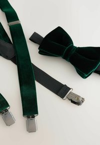 Green velvet bow tie next to green velvet suspenders and black elastic strap. Metal clips and textured fabric details are visible.
