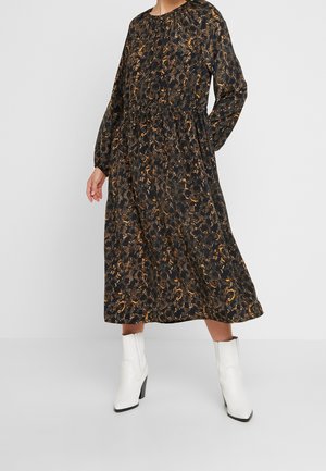 Kobieta w czarno-brązowej, wzorzystej sukience midi z długim rękawem, w białych botkach do kostki, stoi na jednolitym tle.