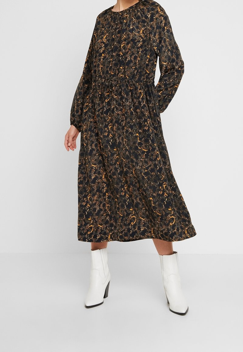 Femme portant une robe midi à manches longues noire et marron à motifs, associée à des bottines blanches, debout devant un fond uni.